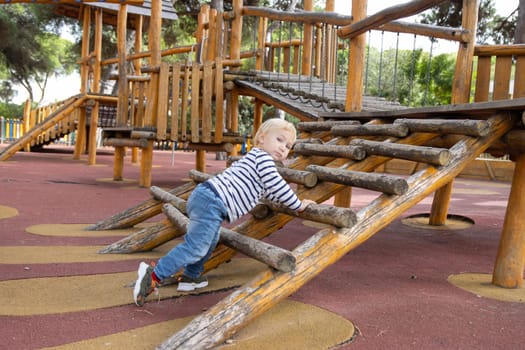 Little blonde boy climbing up wooden bars on the playground - looking at the camera. Mid shot
