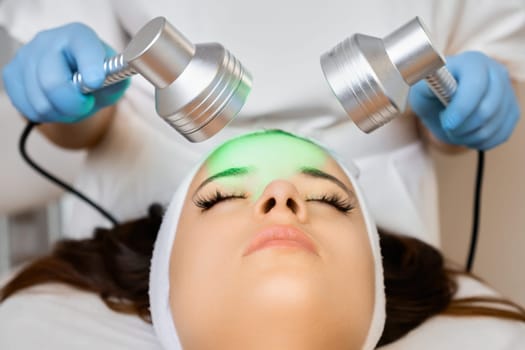 Young attractive woman having light facial treatment with green color in beauty salon.