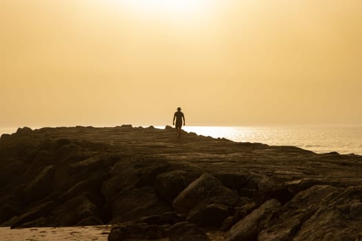 Silhouette of a man wearing wireless headphones walking on a cliff at sunset. Mid shot
