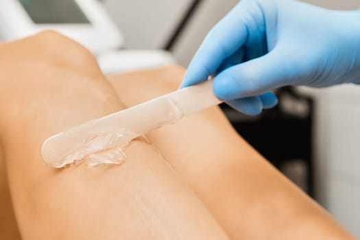 Woman specialist smears wax on stick on female leg in salon light office. Concept of epilation and equipment for procedure by professional