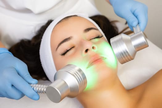 Expert makes light therapy procedure of woman face with contemporary equipment. Concept of procedure to improve female skin condition