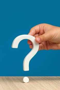 The eternal question, the unknown or the search for an answer. A hand holding a white question mark on a blue background. Vertical photo with copy space. High quality photo