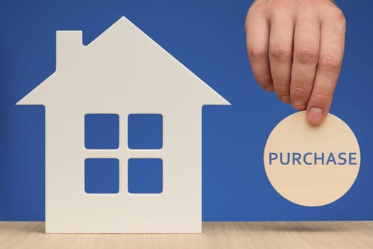 Buying a house. White house model as a symbol of real estate purchase. Close-up hand holding a wooden sign with the inscription PURCHASE. Banner on the theme of real estate with a blue background