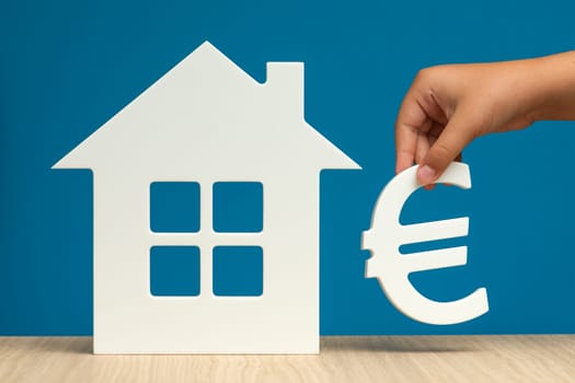 The cost of renting real estate in Europe. Renting residential real estate in the European Union. Model of a white house and a hand with a euro symbol on a blue background close-up. copy space.