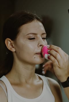 Portrait of a young beautiful Caucasian girl sitting on a chair in a beauty salon, to whom the master is applying foundation cream with a sponge on her face, side view.