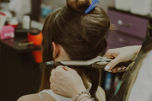 Portrait of a young Caucasian girl sitting in a hairdresser's shop with a master twisting her hair on a flat iron.