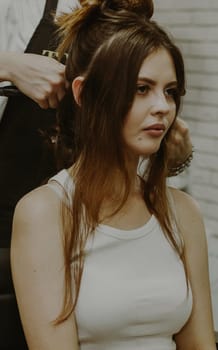 Portrait of a young Caucasian girl sitting in a hairdresser's shop with a master twisting her hair on a flat iron.