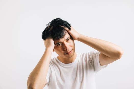 An Asian man, overwhelmed by stress, holds his head in pain with a sad expression. Studio shot isolated on white, conveying the feeling of tension and sickness.