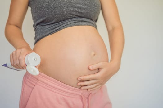 Positive young pregnant woman in comfortable homewear applying belly butter on her big tummy, woman belly.