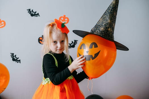 Children's Halloween - a girl in a witch hat and a carnival costume with airy orange and black balloons at home. Ready to celebrate Halloween.