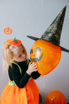 Children's Halloween - a girl in a witch hat and a carnival costume with airy orange and black balloons at home. Ready to celebrate Halloween.