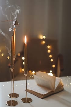 Still life details of home interior, concept of coziness and home atmosphere. beautiful golden candlesticks, scented candle, open book, garland lights. Home autumn concept