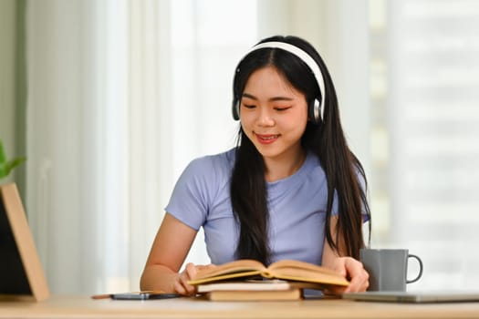 Charming young Asian girl wearing headphone and reading interesting book in bright living room.