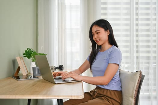 Pleased young asian woman in casual clothes using laptop on wooden desk at home.