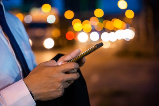 Portrait Asian businessman typing an sms message via smartphone after work near office at night city street, bokeh lights, young man walking and chatting with friends at social networks outdoor