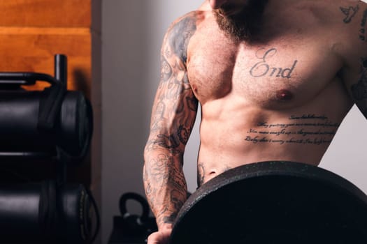 crop of unrecognizable sportsman with muscular body and naked torso standing with heavy weight plate during workout in gym, concept of fitness and active lifestyle