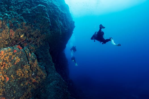 three scuba divers in wetsuit diving near of a wall of rough rocky formation in deep blue sea with clear water, concept of active leisure in ocean and underwater adventure, copyspace for text