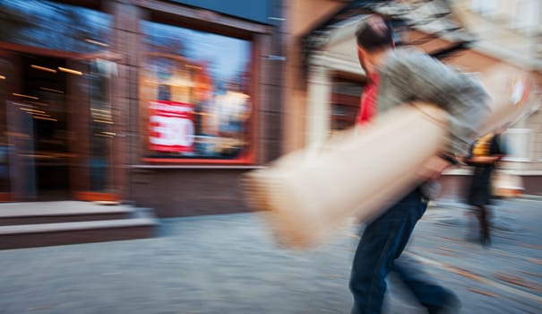 Blurry motion image of a man carrying a large bag, walking past a store with a sale sign in the window.