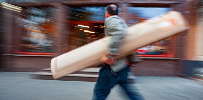 A man carries a long, slender package down a city street, blurred for motion effect.
