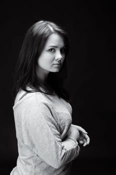 A young woman with long dark hair looks pensively over her shoulder, her arms crossed in front of her in a gray sweater.