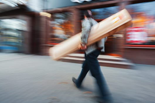 A person carries a long, slender package down a city street, captured with motion blur.