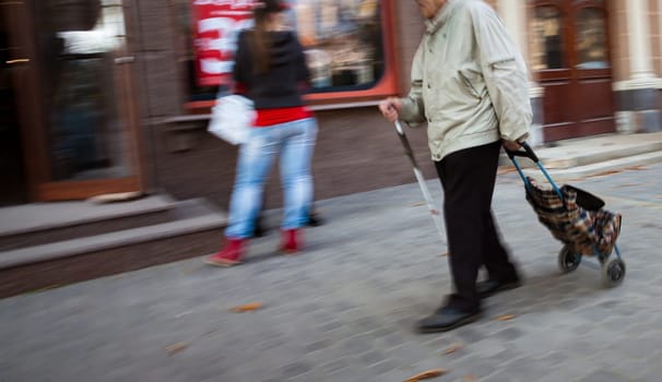 A visually impaired senior citizen with a cane and a wheeled cart walks down a city street, with a blurred woman walking past him.