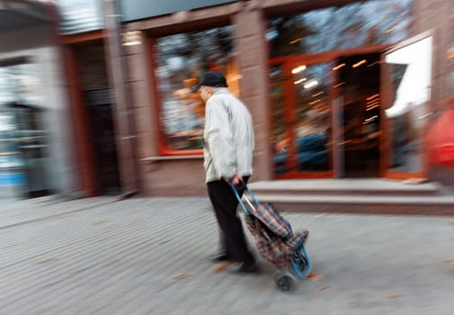 An elderly man with a shopping cart walks briskly down a city street, the background blurred by motion.