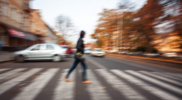 A person walks across a city crosswalk as cars speed by in a motion blur.
