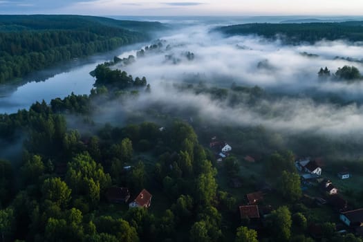Soft fog blankets the serene river valley at dawn, partially obscuring quaint houses nestled among lush green trees.
