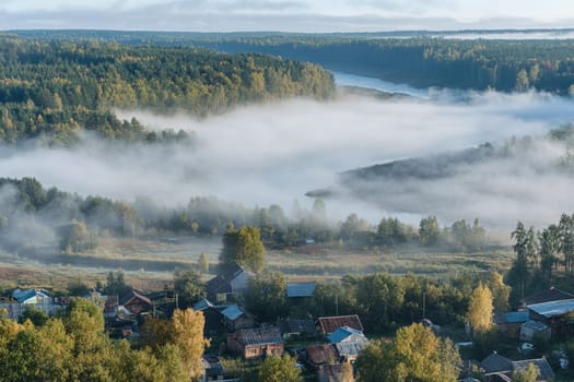 Early morning fog envelops a quiet village along the river, creating a serene atmosphere in the surrounding forested landscape.