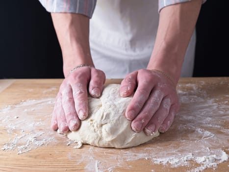 A close up photo of the hands of a man forming dough into a ball on a wooden table.