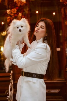 girl standing and holding a cute white dog in her arms. The girl is dressed in a white pantsuit. The dog is also white and fluffy, a Spitz breed.