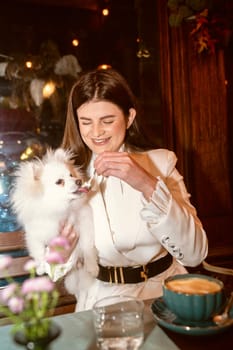 A person sitting at a table with a dog.A girl in a white suit holds a small white dog in her arms. The dog is fluffy, a Spitz breed. The dog is very cute, the girl is smiling.
