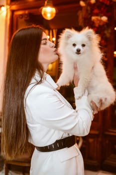 girl standing and holding a cute white dog in her arms. The girl is dressed in a white pantsuit. The dog is also white and fluffy, a Spitz breed.
