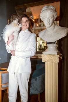 a girl stands in a cafe with a dog in her arms. High quality photo, girl in a white suit, she is tall with long hair. the dog is white, fluffy, spitz breed. the dog licks the girl in the face