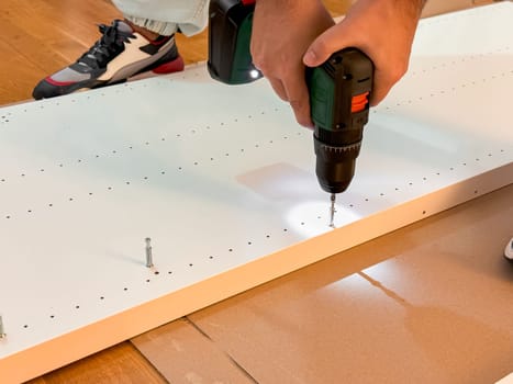 Male assembling furniture using electric drill on wooden board.