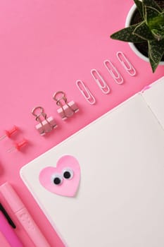 Heart-shaped sticker with eyes glued on the open page of a notepad. Pink themed background with stationery, paper clips, clips, sharpener, notepad, pencil, pen, eraser. Space for your text.