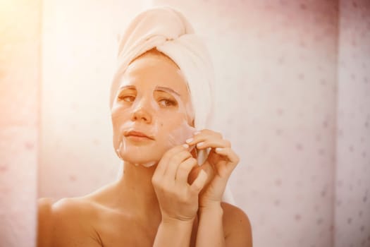 Young beautiful woman using a moisturizing facial mask after taking a bath. Pretty attractive girl in a towel on her head stands in front of a mirror in a home bathroom. Daily hygiene and skin care.