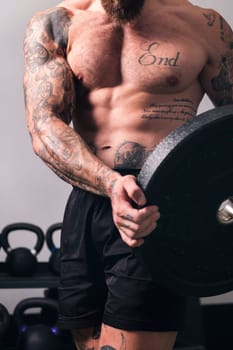 crop of unrecognizable sportsman with muscular body and naked torso standing with heavy weight plate during workout in gym, concept of fitness and active lifestyle