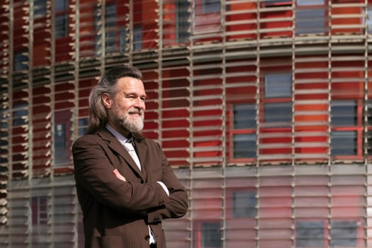 positive middle age male with beard in elegant suit standing with arms crossed and looking away pensive against urban building, concept of business and mature man lifestyle, copyspace for text
