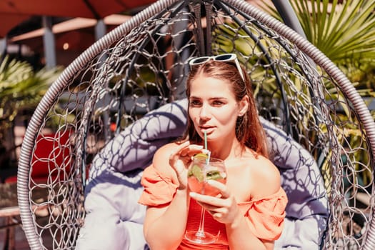A woman in an orange dress is sitting in a hanging chair. She is holding a wine glass and smiling. The scene takes place in a restaurant with several chairs and tables
