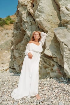 A woman in a white dress is standing on a rocky beach. Concept of serenity and tranquility, as the woman is enjoying the natural beauty of the beach