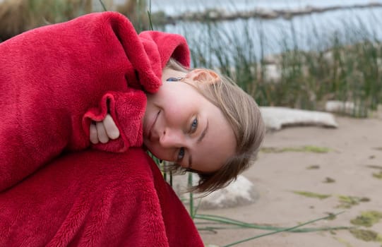 Woman wrapped in a red blanket sits on the beach, looking down and smiling slightly. 