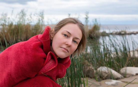 A young woman in a red blanket looks at the camera while crouching near tall grass by the sea.