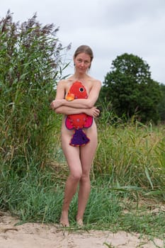 A woman stands in a natural setting, holding a vibrant fish plush toy. The scene is surrounded by tall grasses and trees, capturing a serene atmosphere.