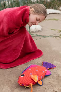 A woman wearing a red blanket leans down to examine a vibrant fish toy resting on sandy ground. The scene captures a tranquil moment by the beach with gentle natural surroundings.