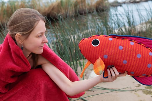 A woman wrapped in a red blanket smiles as she playfully holds a vibrant plush fish at the waters edge. Surrounding grasses and serene water create a peaceful atmosphere.