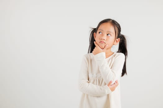 Portrait Asian kid girl deep in thought, standing with chin in hand, pondering a question. Studio shot isolated on white with ample copy space, kindergarten child portraying contemplative expression