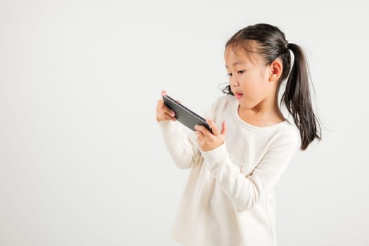 Happy Asian portrait cute young kid girl smiling excited using smart mobile phone studio isolated on white background, Thai kindergarten child surprised holding reading message gesture on smartphone