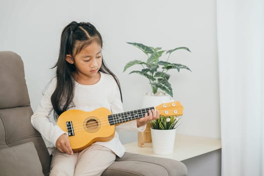Little Asian girl plays a ukulele on a stylish sofa. Dressed in a white shirt, the living room soft tones and decor highlight the peaceful and joyful mood of this musical moment.